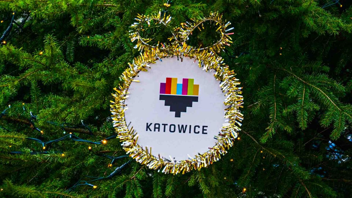 Na gałęziach choinki widać okrągłą ozdobę z napisem „Katowice” i kolorowym logo miasta. Dekoracja jest obramowana złotą lametą i zawieszona na zielonych igłach. W tle widoczne są światełka choinkowe oraz fragmenty przewodu lampek.