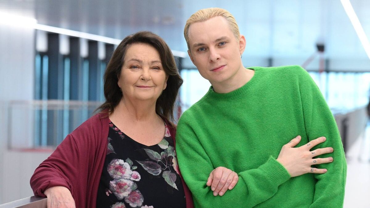 Anna Dymna i Ralph Kamiński stoją obok siebie w jasnym, nowoczesnym wnętrzu, patrząc wprost w obiektyw z łagodnymi uśmiechami. Aktorka ma na sobie kwiecistą sukienkę i bordowy kardigan, a wokalista wyróżnia się intensywnie zielonym swetrem i jasnymi włosami. Ich bliska, swobodna poza podkreśla serdeczną relację i ciepły charakter wspólnego zdjęcia.