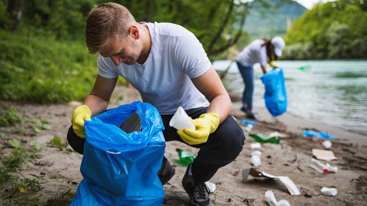Mężczyzna w rękawicach zbiera odpady do niebieskiego worka na brzegu rzeki. W tle widoczna jest druga osoba sprzątająca teren nad wodą. Na ziemi znajdują się plastikowe butelki i inne śmieci.
