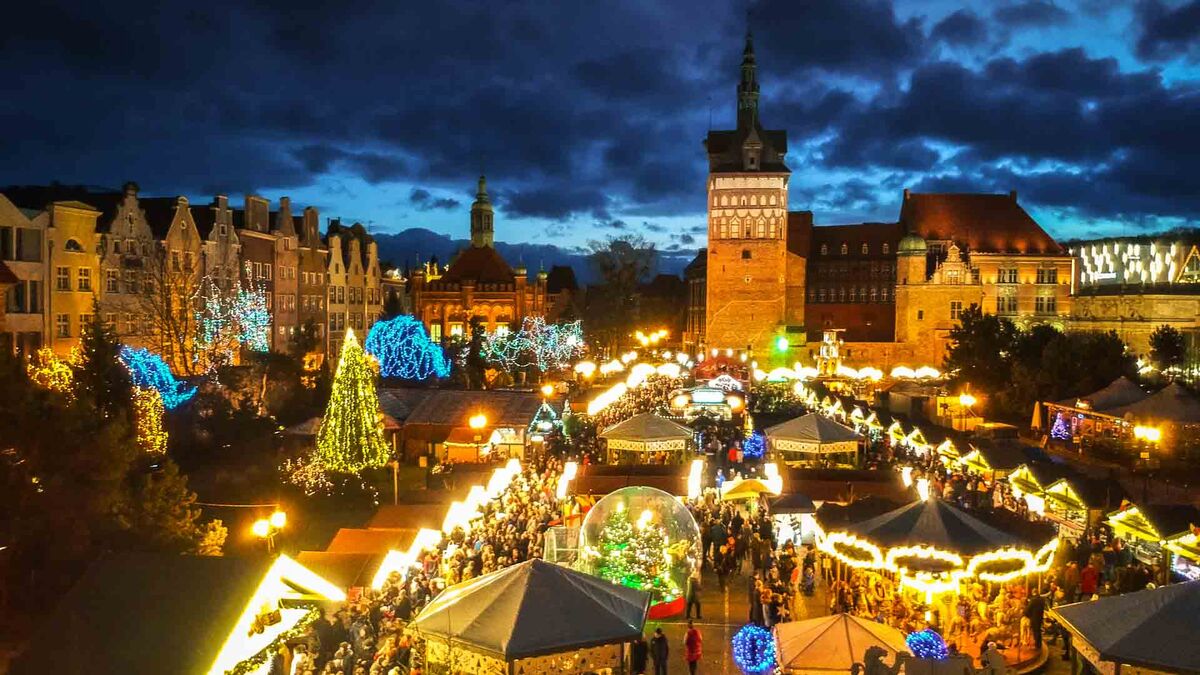 Jarmark Bożonarodzeniowy w Gdańsku. Nocna panorama jarmarku z góry z widocznymi szpalerami stoisk i tłumem na głównych alejach. W tle dominuje sylwetka Bramy Wyżynnej oraz zabudowa śródmieścia. Na terenie jarmarku widać karuzelę, kopułę świetlną i kolorowe instalacje.