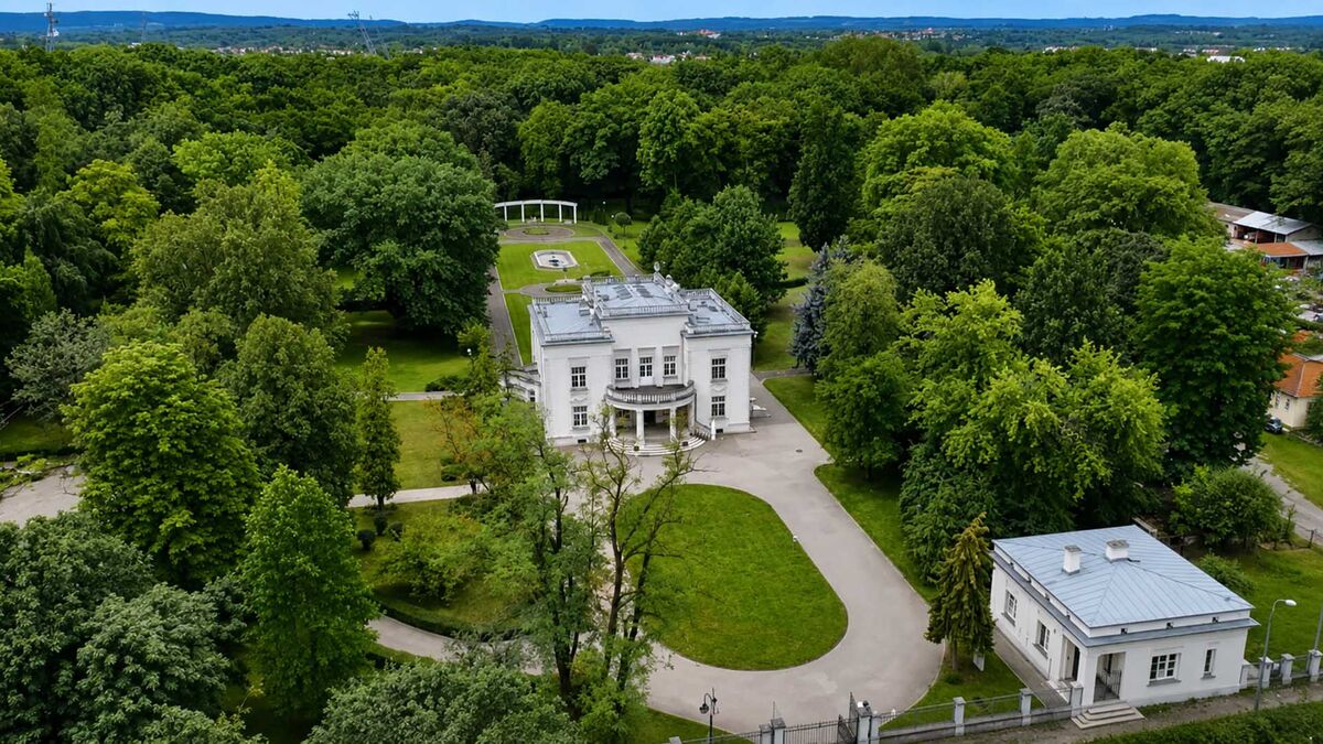 Biały budynek otoczony rozległym parkiem znajduje się w dzielnicy Mościce w Tarnowie. Przed budynkiem prowadzą alejki, a z tyłu rozciąga się uporządkowana część ogrodu. Wokół rosną liczne wysokie drzewa, a w oddali widać zabudowania i wzniesienia.