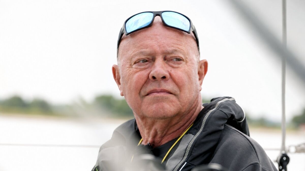 Roman Paszke siedzi na pokładzie jachtu i patrzy w bok. Na głowie ma okulary przeciwsłoneczne oparte na czole, a ubrany jest w ciemną kurtkę. W tle widoczna jest rozmyta tafla wody oraz elementy wyposażenia jachtu.