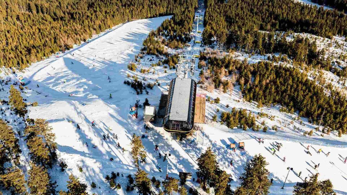 Szeroki, ośnieżony stok narciarski przecina gęsty las iglasty, a po zboczu widać wielu narciarzy i snowboardzistów. W centralnej części kadru znajduje się dolna stacja wyciągu z zadaszeniem, od której w górę prowadzi linia krzesełek. Długie cienie drzew i ludzi padają na śnieg, co sugeruje niskie słońce.