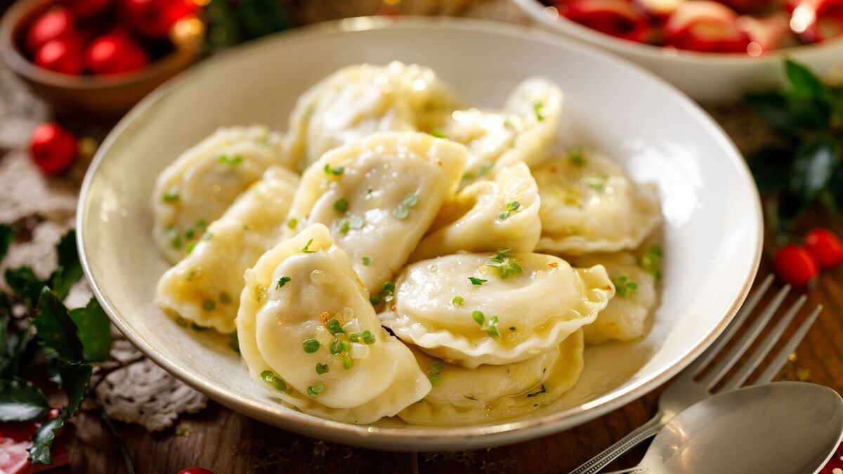 W jasnej misce leży porcja pierogów z sandaczem, polanych roztopionym masłem i posypanych drobno siekanymi ziołami. Pierogi mają złocisty połysk i są ułożone warstwami, częściowo zachodząc na siebie. W tle widać rozmyte świąteczne dekoracje w czerwieni oraz sztućce przy brzegu stołu.