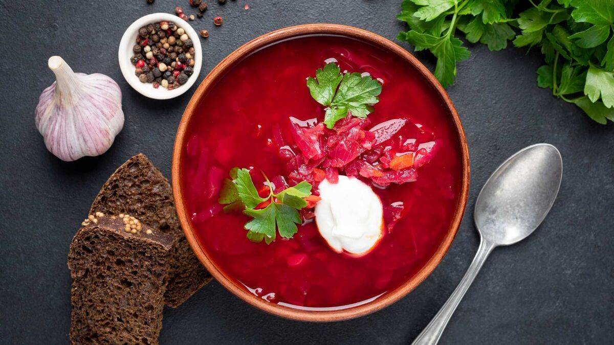 Aromatyczna zupa z botwinki podana w misce zachwyca intensywnie czerwonym kolorem. W środku widać kawałki warzyw, a całość udekorowana jest śmietaną i natką pietruszki. Obok znajdują się kromki ciemnego chleba, czosnek, pieprz w ziarnach oraz łyżka, tworząc domową kompozycję.
