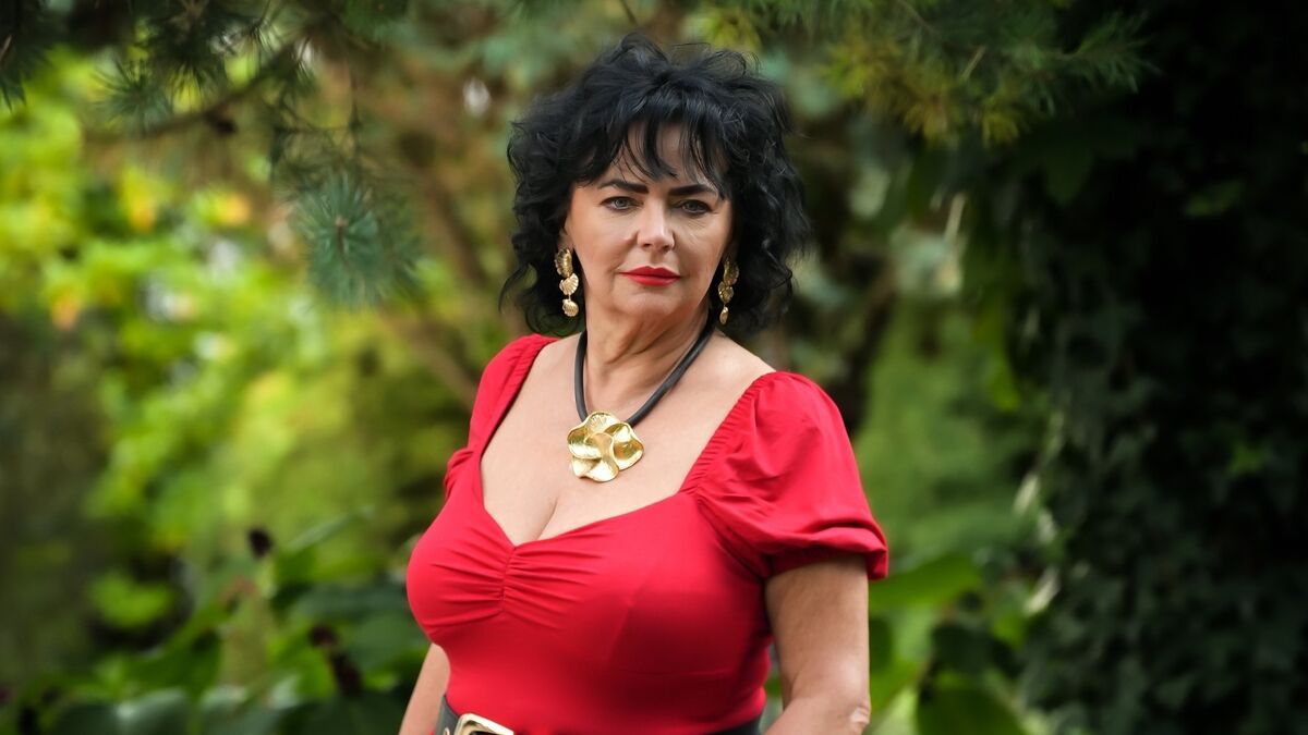 Emilia ma na sobie czerwoną sukienkę z krótkimi bufiastymi rękawami oraz szerokim dekoltem. Jej stylizację uzupełniają duży złoty naszyjnik i wiszące kolczyki w podobnym kolorze. W tle widoczna jest gęsta zielona roślinność, która stanowi naturalne otoczenie.