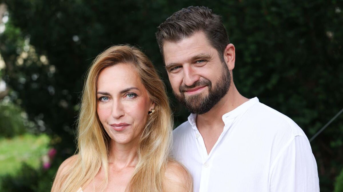 Magdalena Waligórska-Lisiecka i jej mąż Mateusz pozują razem, stojąc obok siebie. Magdalena ma długie blond włosy i złote kolczyki, a Mateusz krótkie ciemne włosy, brodę oraz białą koszulę. W tle widoczna jest gęsta zielona roślinność oraz fragment trawnika.