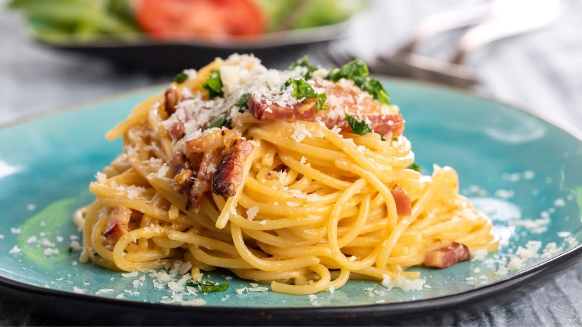 Na turkusowym talerzu ułożona jest porcja spaghetti carbonara z wyraźnie widocznymi kawałkami podsmażonego boczku. Makaron pokrywa kremowy sos, a całość posypano drobno startym serem i świeżą natką pietruszki. W tle, poza ostrością, widać fragmenty zastawy stołowej i misę z warzywami.