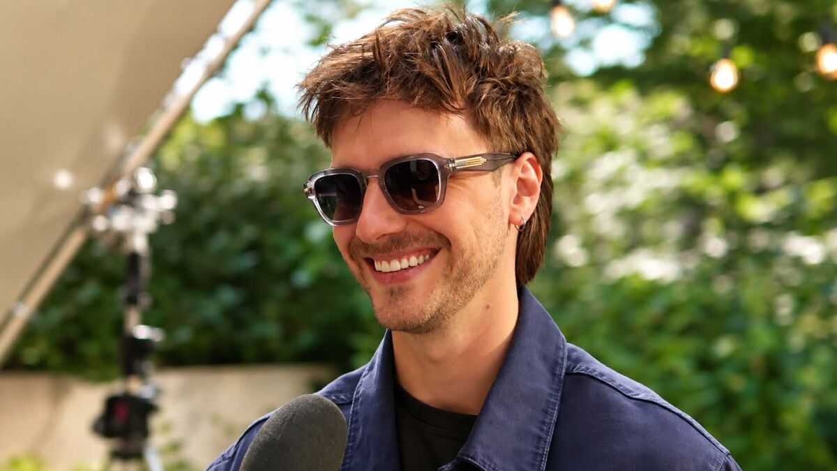 Maciej Musiał stoi na zewnątrz w ciemnych okularach przeciwsłonecznych i granatowej kurtce. Przed nim widoczny jest mikrofon, a mężczyzna ma krótkie, uniesione włosy i niewielki kolczyk w uchu. W tle znajdują się drzewa, zielona roślinność oraz elementy wyposażenia planu.
